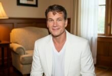Headshot of a smiling man in a white suit jacket against an interior background with soft lighting, suggesting a professional or relaxed setting.